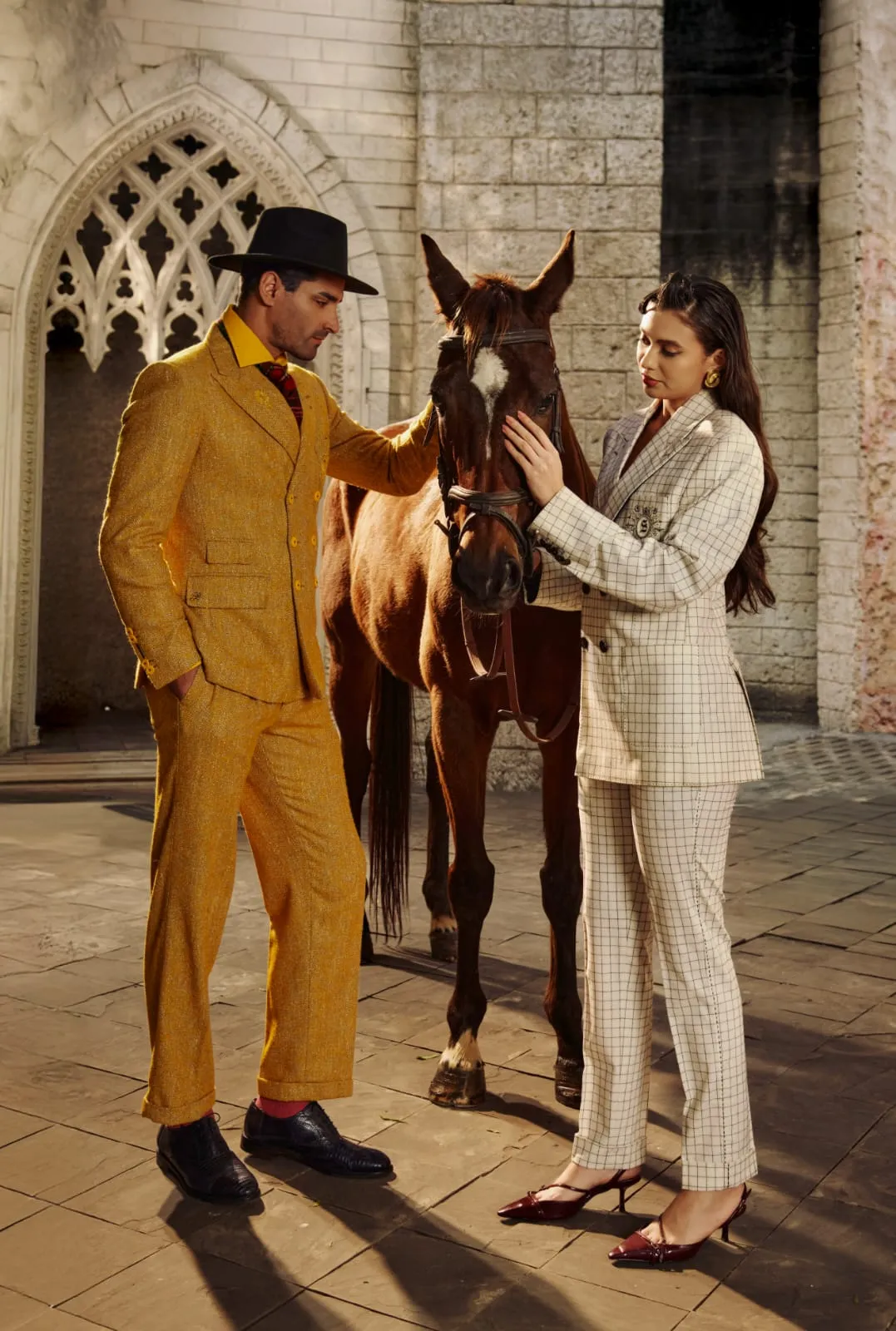 A couple in a yellow suit and white pantsuit beside a chestnut horse, gothic stone arch behind.
