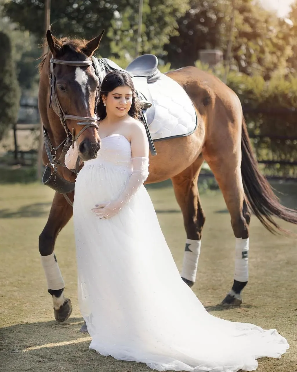 A woman in a pearl-trimmed white dress beside a chestnut horse in golden light.