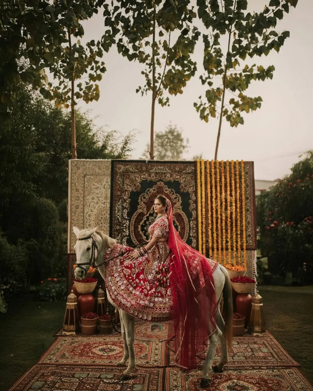 A bride in red lehenga sidesaddle on a white horse, before a tapestry-and-marigold backdrop on the lawn.
