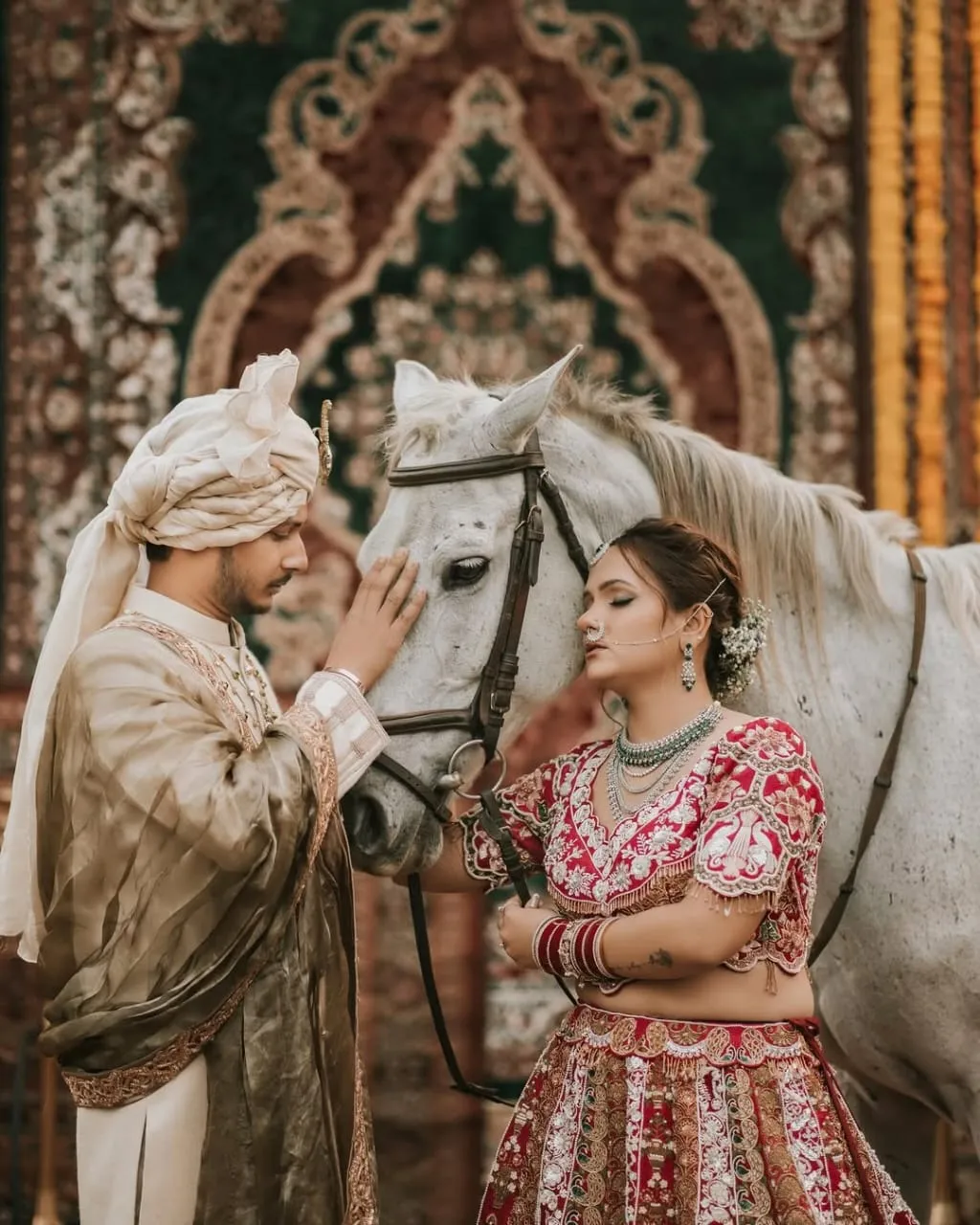 A groom in a sherwani seated, bride sidesaddle on a dark horse, against a richly patterned tapestry backdrop.